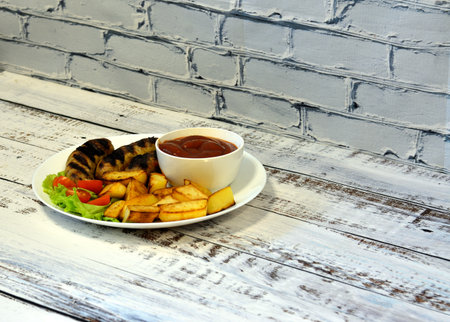 Grilled sausages with fried potatoes and tomatoes on a large plate with a cup of sauce and a leaf of lettuce on a light wooden table. Top view, flat lay.の写真素材