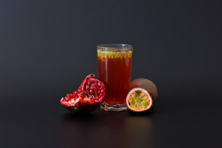 A tall glass of fruit juice with seeds on a black background, next to pieces of ripe passion fruit and a pomegranate fruit. Close-up.の写真素材