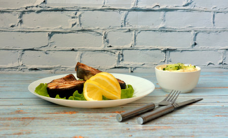 Two grilled pink salmon steaks with lettuce and lemon slices on a wooden table, cutlery and a cup with sauce nearby. Close-up.の写真素材
