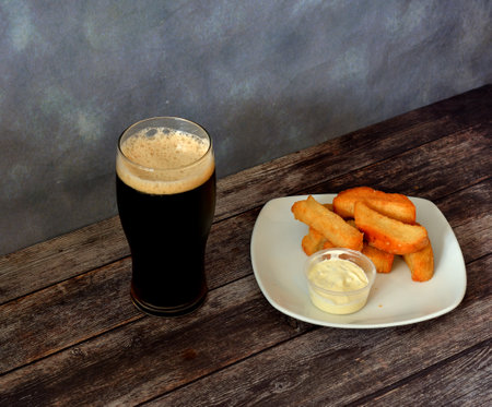 A plate of wheat croutons with sauce and a tall glass of dark beer with white foam on a dark wooden...