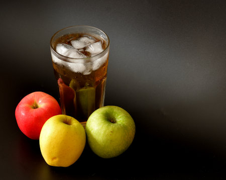 Apple juice with ice in a tall faceted glass on a black background, with ripe fruits nearby. Close-up.の写真素材
