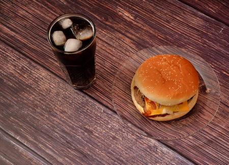 A glass plate with a large hamburger with meat cutlet and sauce and a tall glass of cola with ice on a dark wooden table. Top view, flat lay.の写真素材