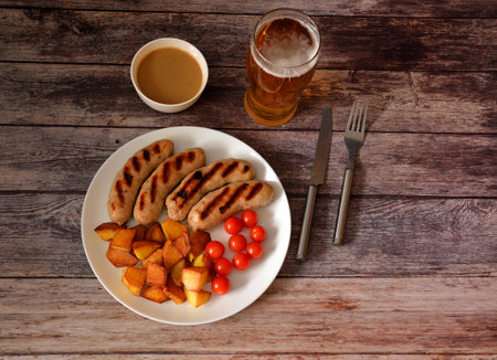 A plate with grilled sausages, cherry tomatoes and fried potatoes on a wooden table, next to a glass of light beer, mustard and cutlery. Top view, flat lay.の写真素材
