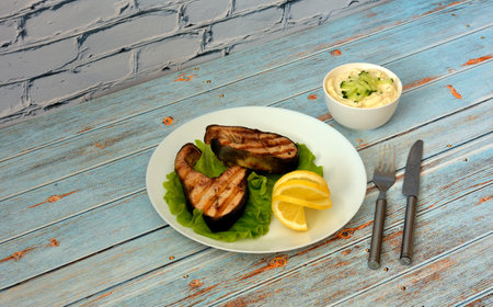 A plate with two grilled salmon steaks with lemon and lettuce on a wooden table, sauce and cutlery nearby. Close-up.の写真素材