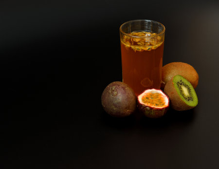 A tall glass of freshly squeezed juice with seeds on a black background, next to a ripe kiwi fruit and pieces of chopped passion fruit. Close-up.の写真素材