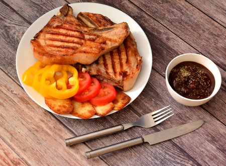 Two fried pork chops with slices of tomatoes, sweet peppers and baked potatoes on a wooden table, cutlery and a cup of sauce nearby. Top view, flat lay.の写真素材