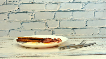 A plate with two fried eggs, crispy bacon slices and bread toast on a light wooden table, cutlery...