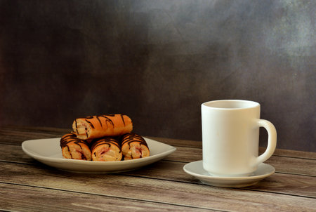 A plate with several mini-rolls in chocolate coating and a cup of hot drink on a saucer on a wooden table. Close-up.の写真素材