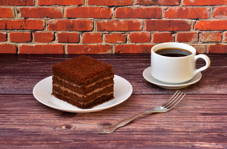 A piece of sponge chocolate layer cake on a white plate and a cup of hot black coffee on a wooden...