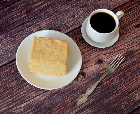 A piece of sponge layered cake with protein cream Napoleon on a white plate and a cup of hot black...