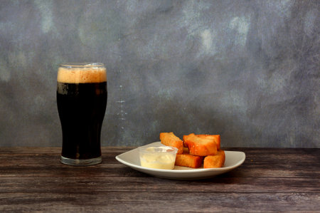 A glass of dark beer with high white foam and a plate with wheat croutons and garlic sauce on a wooden table. Close-up.の写真素材