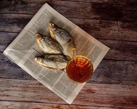 A mug of light beer on a newspaper and three dried river fish on a dark wooden table. Top view, flat lay.の写真素材
