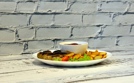 Plate with grilled sausages, fried potatoes and tomato sauce on a light wooden table. Close-up.
