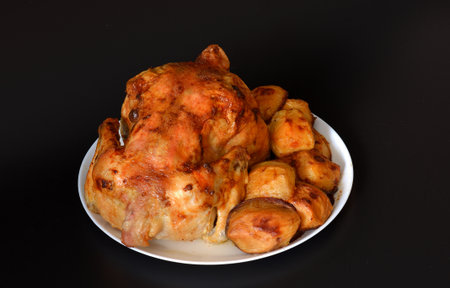 Whole baked chicken with new potatoes on a white ceramic plate on a black background. Close-up.の写真素材