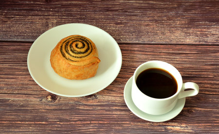 Plate with a spiral poppy seed bun on a wooden table, a cup of hot black coffee next to it. Close-up.の写真素材