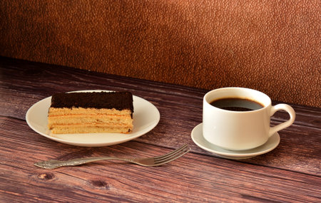 A piece of layered sponge cake with chocolate sprinkles, a fork and a cup of hot black coffee on a wooden table. Close-up.の写真素材