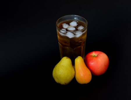 Apple and pear juice with ice in a tall glass on a black background, ripe fruits of different varieties nearby. Close-up.の写真素材