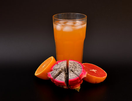 Fruit juice from orange, grapefruit and dragon fruit in a tall glass with ice on a black background, pieces of ripe fruits nearby. Close-up.の写真素材