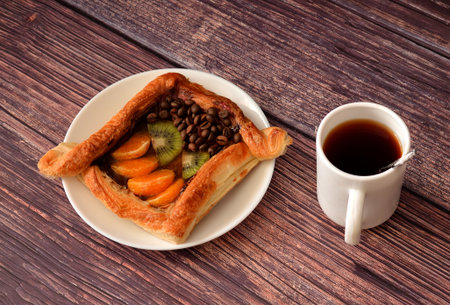 Freshly baked puff pastry bun with kiwi and tangerine pieces and decorated with coffee beans on a wooden table, next to it a cup of hot coffee. Top view, flat lay.の写真素材