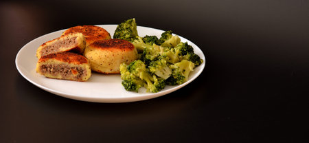 Potato patties stuffed with minced meat and boiled broccoli in a round plate against a black...