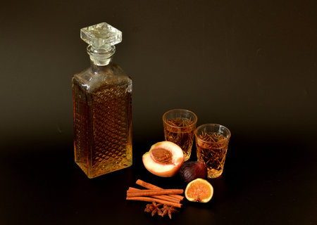Fig-peach liqueur with cinnamon and anise on a black background, a crystal decanter and two glasses with strong alcohol next to spices and and ripe fruit. Close-up.の写真素材