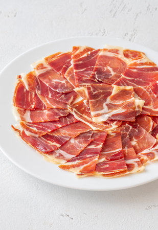 Cured Spanish ham slices fan out delicately across clean, white ceramic round plateの写真素材