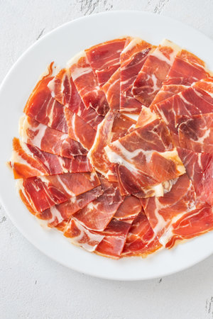 Cured Spanish ham slices fan out delicately across clean, white ceramic round plateの写真素材