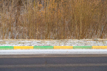 Multicolored stones of the border and a lawn covered with snow next to the roadway against the background of bushes, minimalismの写真素材