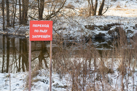 A small river, along the banks of dry grass and trees covered with snow, a shield with an inscription in Cyrillic, crossing the ice is prohibitedの写真素材