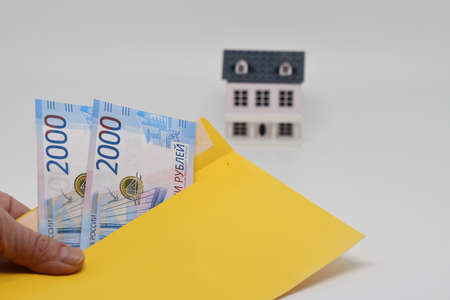 A hand holds a yellow envelope with banknotes against the background of a toy house, funds for the purchase of real estate, mortgageの写真素材