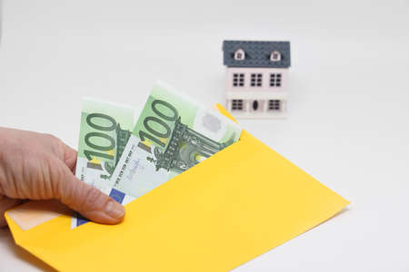 A hand holds a yellow envelope with banknotes against the background of a toy house, funds for the purchase of real estate, mortgageの写真素材