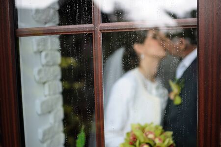 Newlyweds kissing behind a window with raindrops.の写真素材