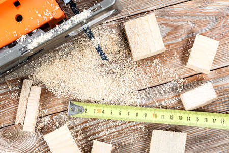 Electric jigsaw with many wooden bricks full of sawdust. On old scratched wooden table, work tools...