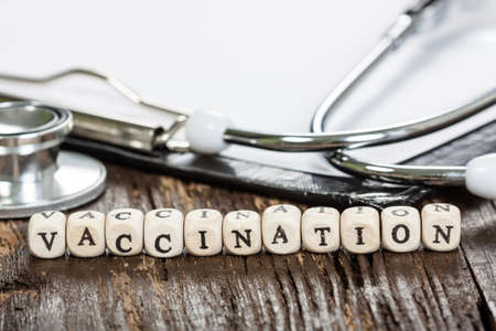 Vaccination word formed by wood alphabet blocks on old dark wooden table, medical clipboard and stethoscope on background. Health care and medicine concept.の写真素材