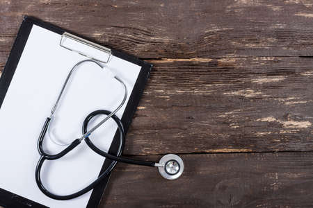 Medical clipboard and stethoscope on old dark wooden table background. Top view. Health care and...