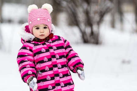 Portrait of little girl in pink jacket with red scarf and pink hat in snowy park at winter.の写真素材