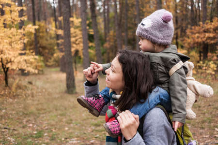 Little girl in autumn forest with teddy bear backpack sitting on mother shoulders. Fall day. Little girl exploring nature with mother.の写真素材