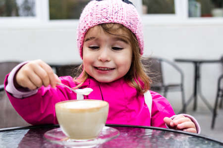 Portrait of a cheerful little girl sitting on the street in city cafe with mom and fooling around with coffee.の写真素材
