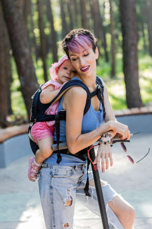 Cool attractive woman with pink hair, enjoys riding an electric scooter with her daughter in sling. Eco friendly transport, electric scooter city transport.の写真素材