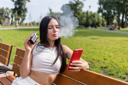 Attractive woman smoking vape device, reading news in the phone at the park.の写真素材