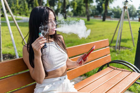 Attractive woman smoking vape device, reading news in the phone at the park.の写真素材