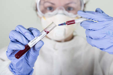 Doctor in protective wear holding test tube with blood for Coronavirus COVID-19 analyzing. Vaccine...