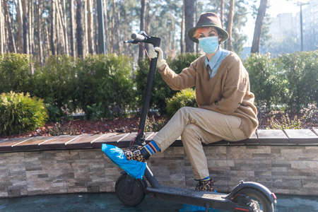 A stylish woman wearing mask and gloves to defened her from coronavirus. Using electric scooter in...