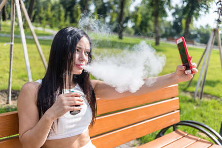 Attractive woman smoking vape device, reading news in the phone at the park.