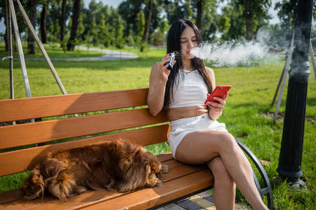 Attractive woman smoking vape device, reading news in the phone at the park.の写真素材