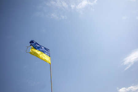 Damaged Ukrainian flag over blue sky during war with russia.