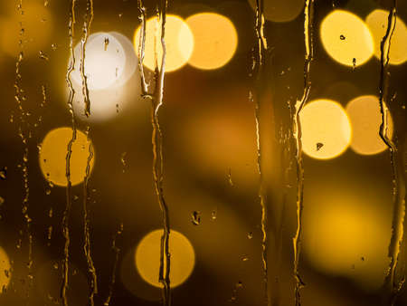 Raindrops and water trickling down a window pane with defocused street lights in the backの写真素材