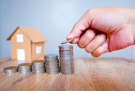 Five of coins stack with human hand on wooden table and house model against gray background for saving and investment real estate concept.の写真素材