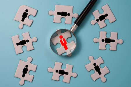 Hand holding magnifier glass to searching red human icon on jigsaw puzzle among black people icon. Human development concept.の写真素材