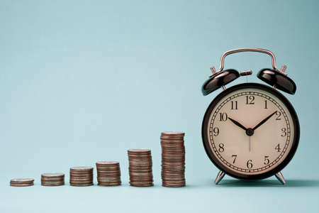 Increase coins stack graph with alarm clock on blue background. Business investment time concept.の写真素材